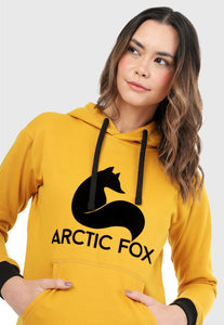 Detalle de Mujer vistiendo la sudadera para mujer conjunto buzo hoodie color mostaza con capota forrada en algodón perchado negro con el zorro de Arctic Fox estampado en terciopelo negro al frente y pantalón de sudadera negro con logotipo en color mostaza.