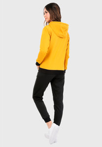 Mujer vista de espalda vistiendo la sudadera para mujer conjunto buzo hoodie color mostaza con capota forrada en algodón perchado negro y pantalón de sudadera negro con logotipo en color mostaza.