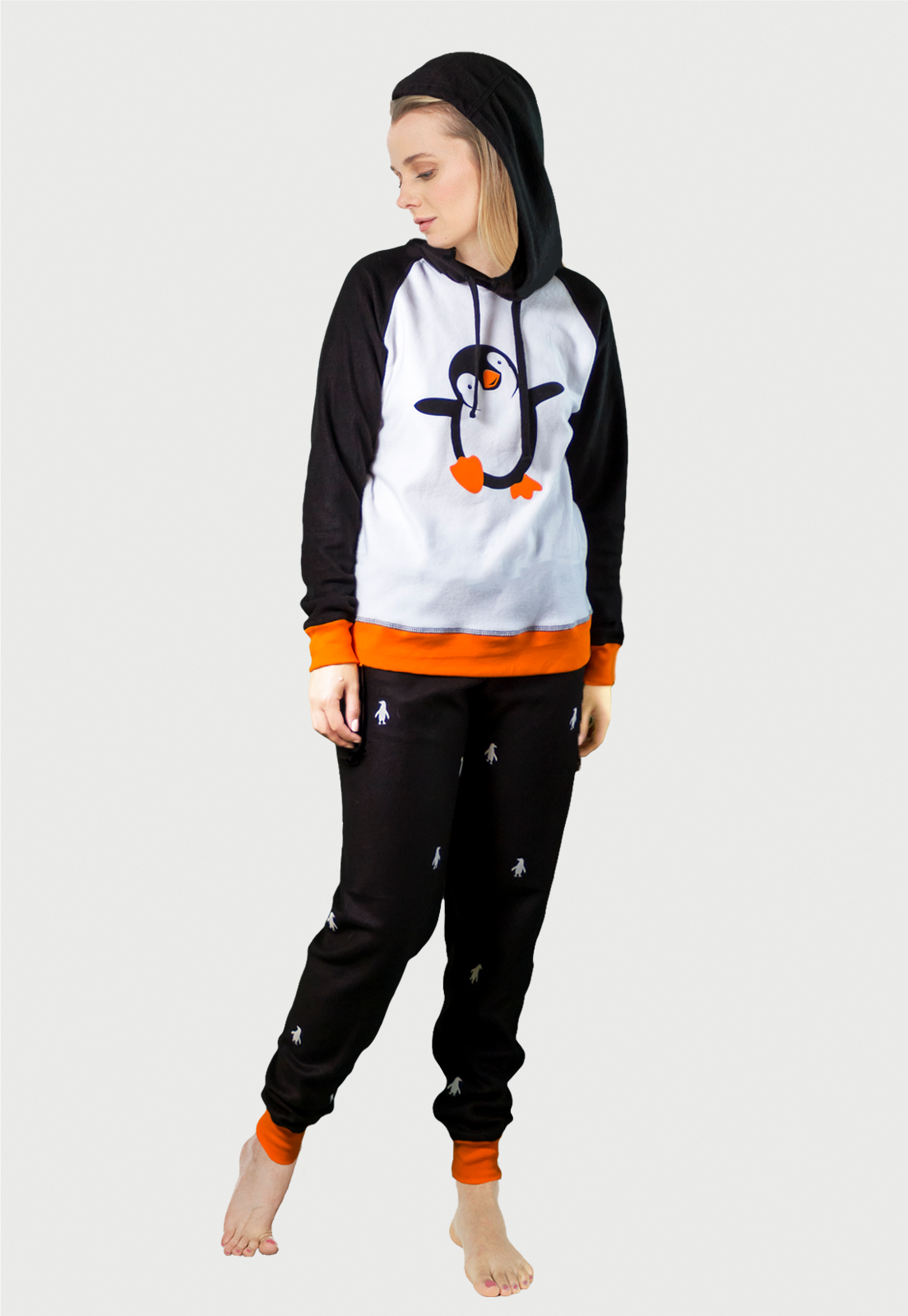 Mujer vistiendo la Pijama para Mujer referencia pingüino con siluetas de pingüino blancas estampadas en el pantalón. El buzo es tipo hoodie. La pijama es de color negro, blanco y naranja con el pingüino bailarín estampado en el pecho, diseño de Arctic Fox Colombia