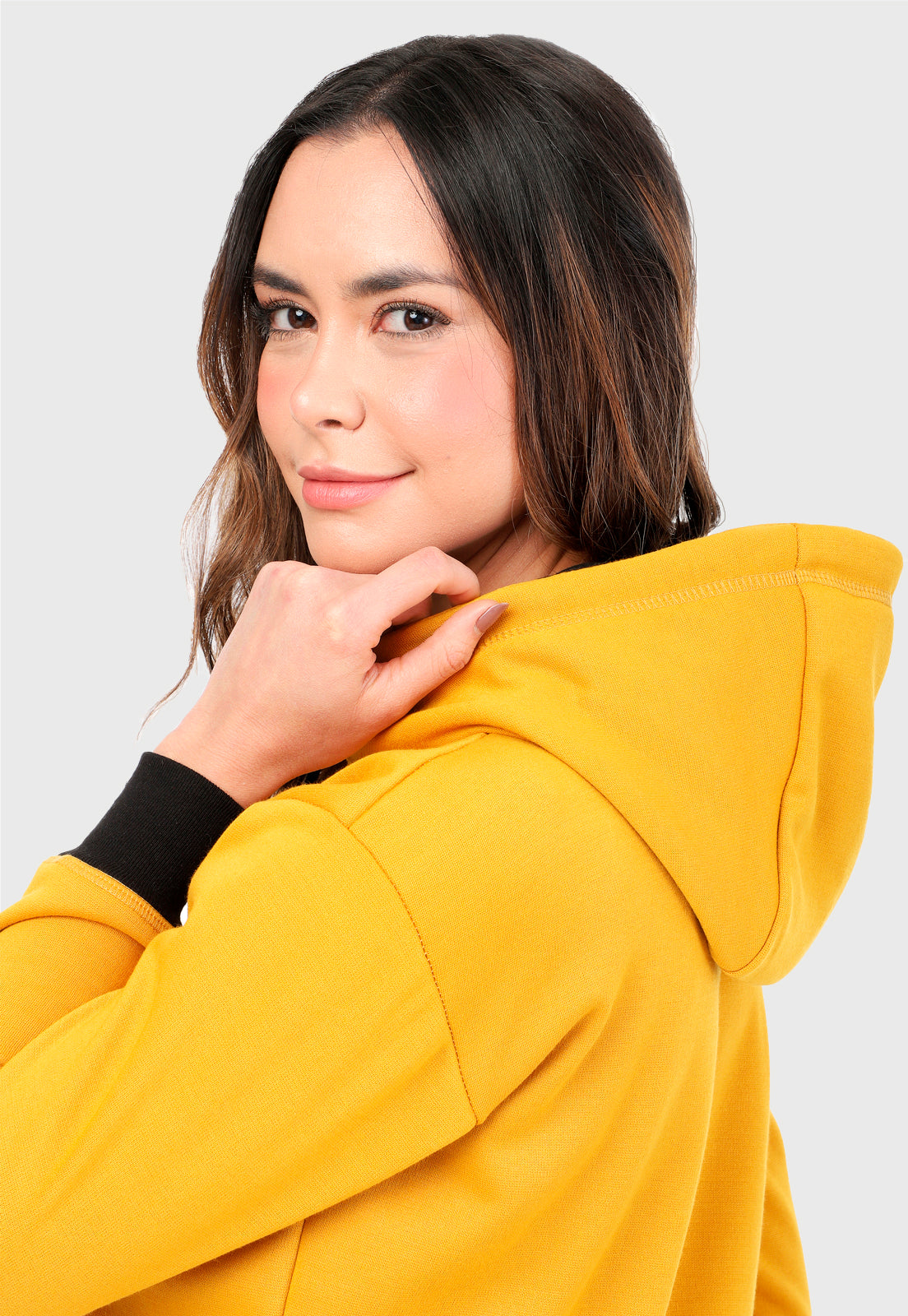Detalle de Mujer vista de lado vistiendo la sudadera para mujer conjunto buzo hoodie color mostaza con capota forrada en algodón perchado negro con el zorro de Arctic Fox estampado en terciopelo negro al frente y pantalón de sudadera negro con logotipo en color mostaza.