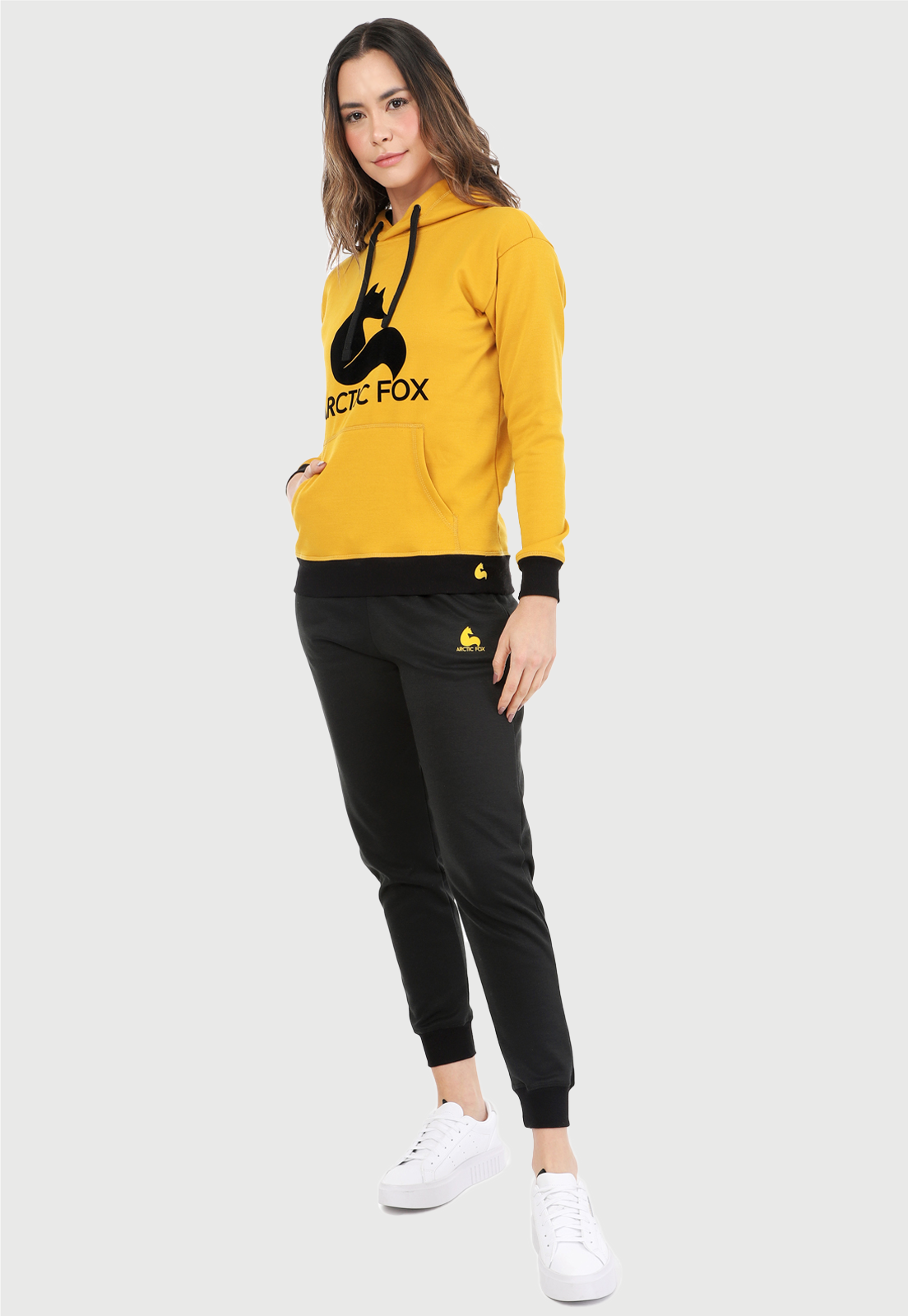 Mujer vistiendo la sudadera para mujer conjunto buzo hoodie color mostaza con capota forrada en algodón perchado negro con el zorro de Arctic Fox estampado en terciopelo negro al frente y pantalón de sudadera negro con logotipo en color mostaza.