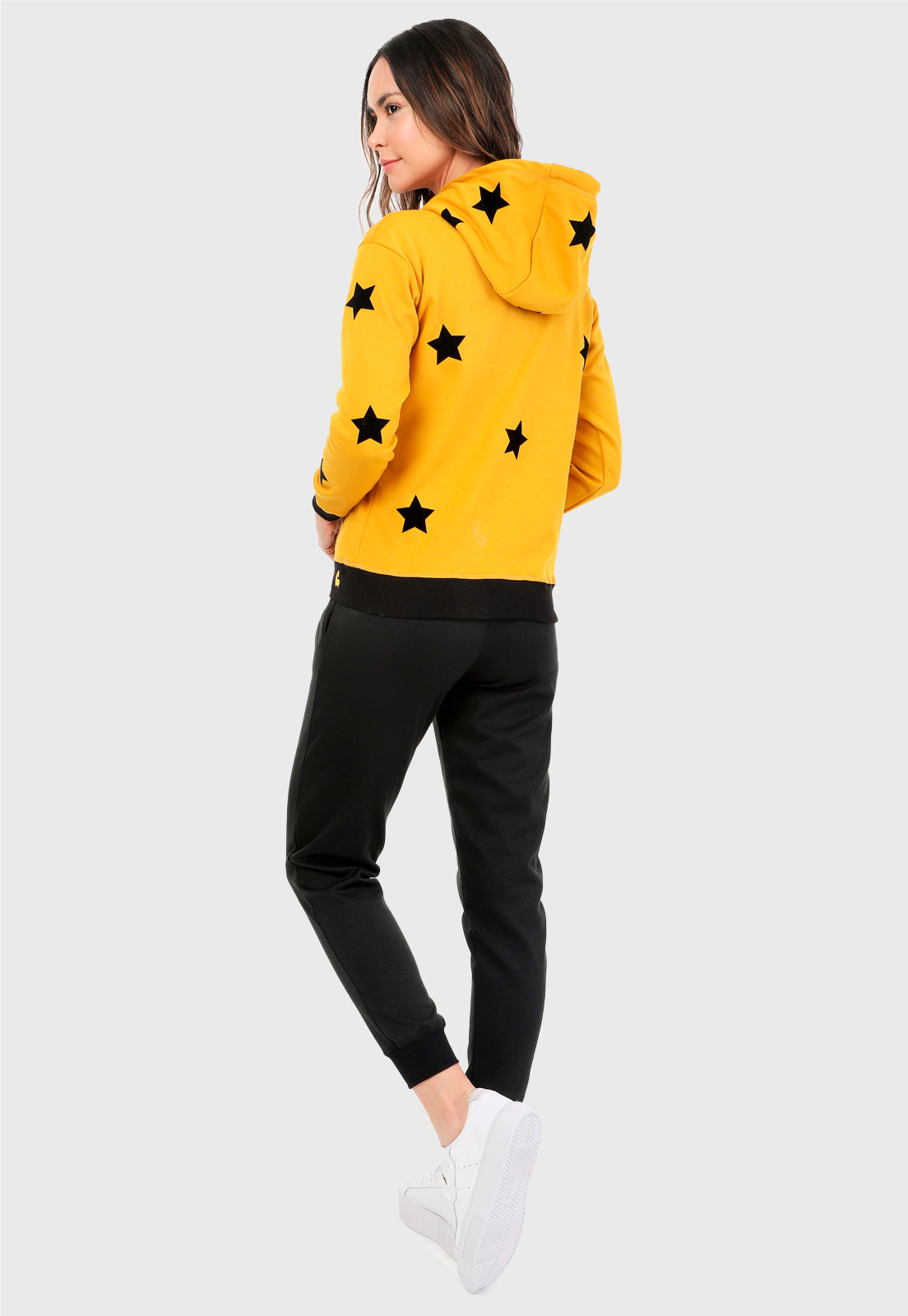 Mujer vista de espalda vistiendo la sudadera para mujer conjunto buzo hoodie color mostaza con estrellas estampadas en negro y pantalón de sudadera negro.