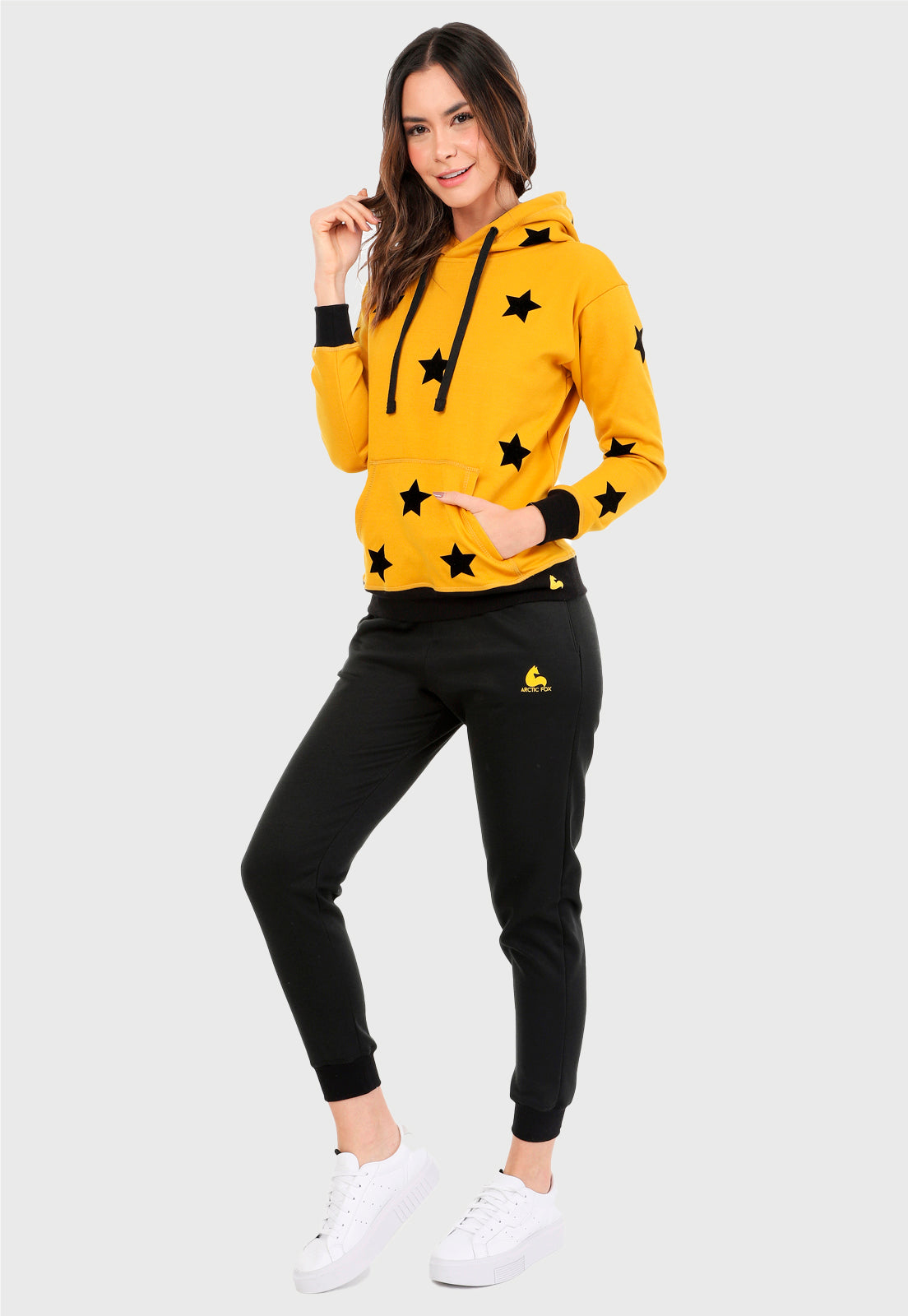 Mujer vistiendo la sudadera para mujer conjunto buzo hoodie color mostaza con estrellas estampadas en negro y pantalón de sudadera negro.